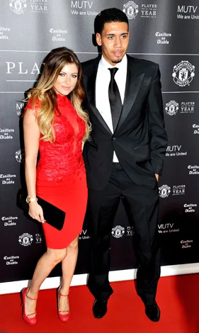Man in a black suit and woman in a red dress pose on red carpet at a Manchester United event backdrop.