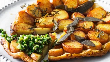 Potato tart topped with herbs and a slice garnished with chopped greens on a white plate.