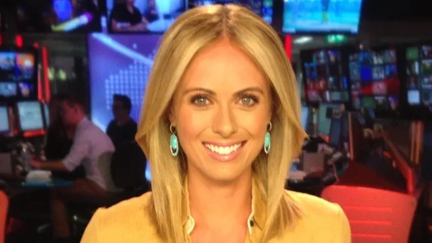 News presenter smiling in a broadcast studio with multiple screens and vibrant lighting.