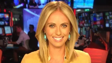 News presenter smiling in a broadcast studio with multiple screens and vibrant lighting.