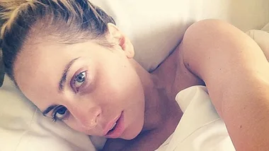 Singer Lady Gaga is lying in bed, makeup-free, looking at the camera with soft lighting.