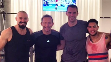"Four men in workout attire posing in a gym, with exercise equipment and a TV in the background."