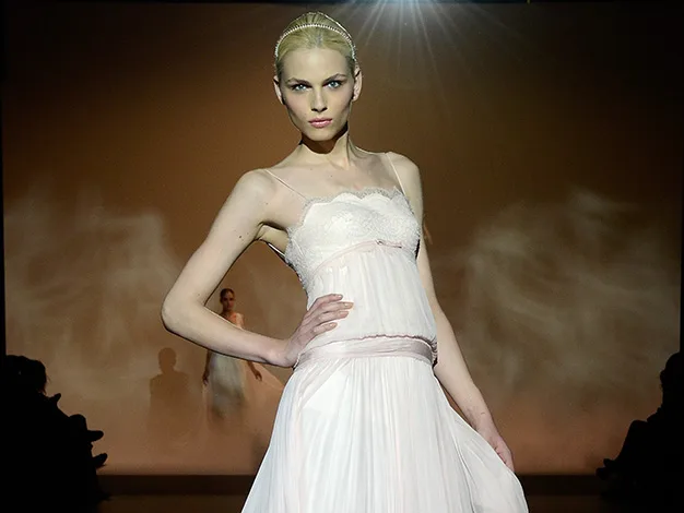 Model on runway in a light pink dress, illuminated by spotlight, with an audience in shadowed seating.