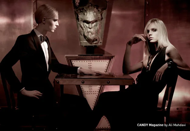 Two androgynous models in formal attire posing at a table in a vintage-style room, photographed by Ali Mahdavi.