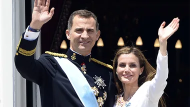King and queen of Spain waving to a crowd from a balcony during a public event.