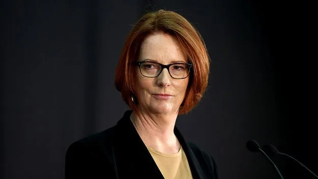 A woman with red hair and glasses speaks at a podium against a dark background.