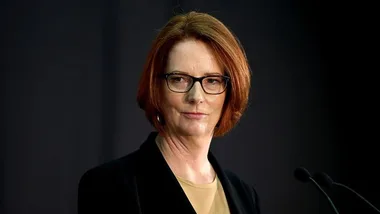 A woman with red hair and glasses speaks at a podium against a dark background.