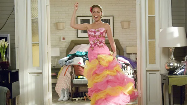 Woman in colorful bridesmaid dress joyfully stands in a room with a bed and lamp.