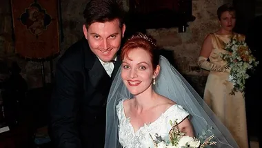 A bride and groom smiling at their wedding, with a bridesmaid holding flowers in the background.