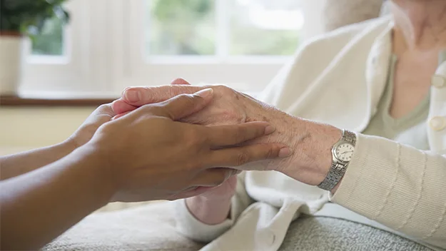 Elderly woman's hands being gently held by a caregiver in a supportive setting.