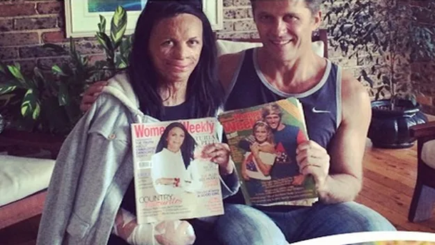 A couple sits on a couch holding magazines featuring them on the cover, smiling at the camera.