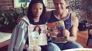 A couple sits on a couch holding magazines featuring them on the cover, smiling at the camera.