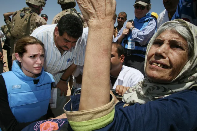 Aid worker in blue vest with refugees and military, an elderly woman gesturing, likely in a Middle Eastern setting.