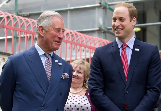 Two men in suits smiling, standing outdoors near red railings, with a woman in the background.