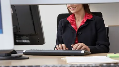 A woman in a blazer types on a computer keyboard in an office setting.