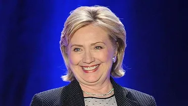 A woman smiling on stage against a blue background, wearing a dark blazer and polka dot top.