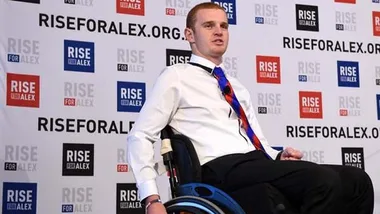 Alex McKinnon in a wheelchair at a "Rise for Alex" event, with related banners in the background.