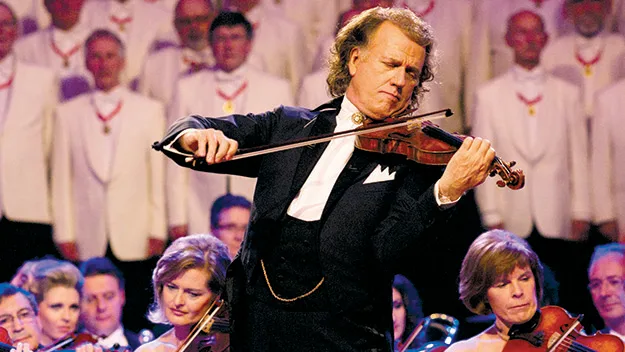 André Rieu playing violin on stage in front of an orchestra and choir, dressed in formal concert attire.