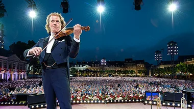 André Rieu plays violin on stage with a large crowd and bright lights in the background at a concert event.