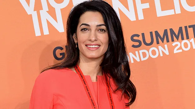 Amal Alamuddin in a coral dress at London Summit 2014, standing against an orange backdrop with white text.