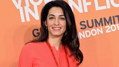 Amal Alamuddin in a coral dress at London Summit 2014, standing against an orange backdrop with white text.