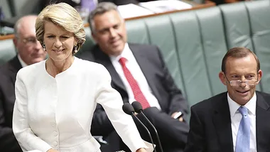 A woman in a white outfit speaks at a podium in a government setting, with men seated around her, one with glasses.