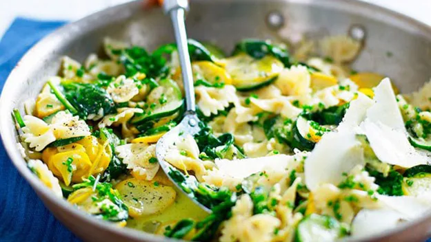 Pasta with zucchini, spinach, and shaved cheese in a skillet, garnished with herbs and black pepper.