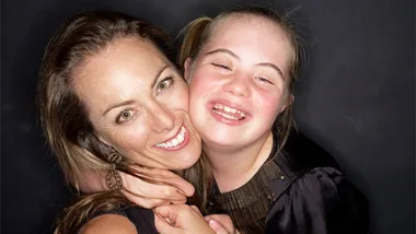 A smiling woman hugs a young girl with Down syndrome, both looking joyful against a dark background.