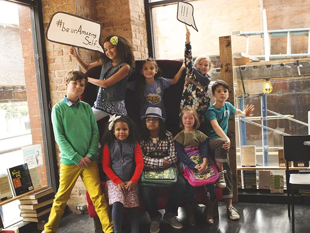 A group of children pose in a cozy setting, holding signs with messages "be ur Amazing Self" and "!!!".