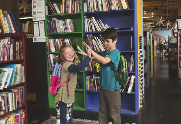 Two children with backpacks high-five joyfully in a library, surrounded by colorful bookshelves.