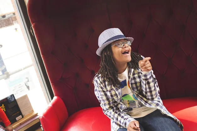 Young person with dreadlocks, wearing a plaid shirt and hat, smiling and pointing while sitting on a red couch.