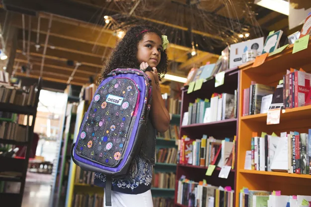 Girl with curly hair holding a decorated backpack stands in a bookstore, surrounded by colorful shelves of books.