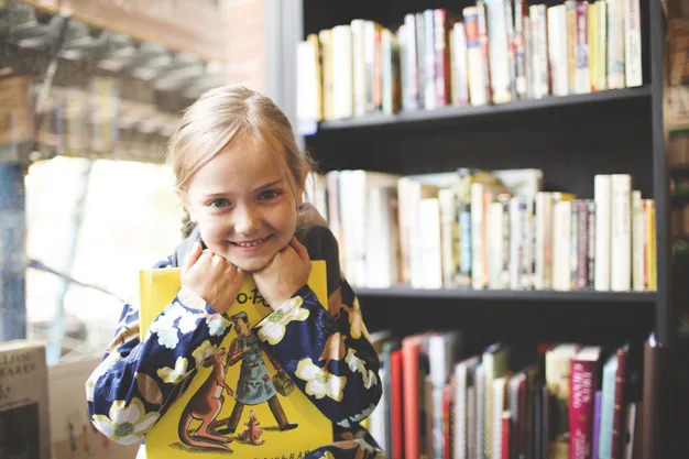 Girl smiling, hugging a yellow book in a library, shelves filled with books in the background.