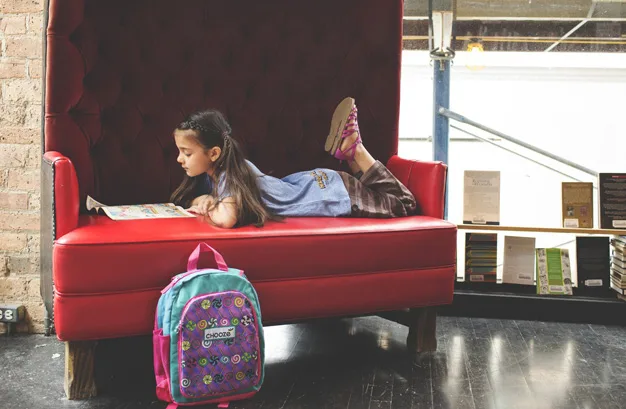 Girl reading a book while lying on a red couch, with a colorful backpack on the floor beside her.