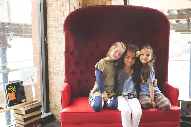 Three young girls sitting on a large red chair, smiling, with books stacked nearby in a cozy setting.