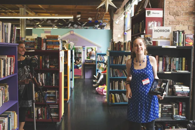 Young woman in a blue dress standing in a library aisle, smiling and holding a book, surrounded by bookshelves.
