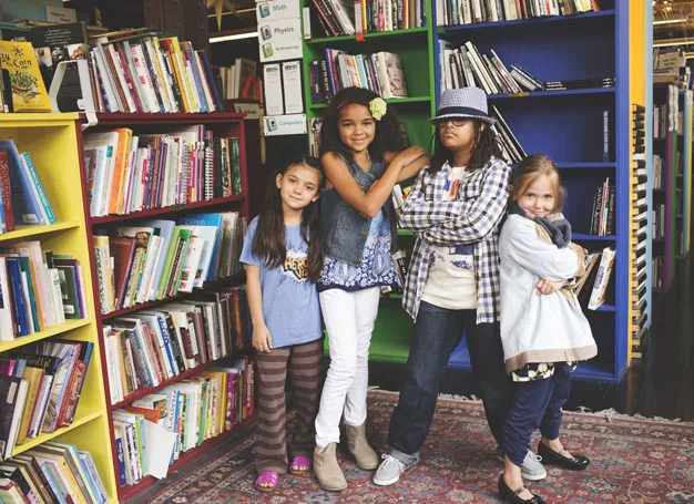 Four children posing confidently in a colorful library, standing next to shelves filled with books.