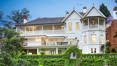 White mansion with wraparound porch, surrounded by lush greenery and trees, under a twilight sky.