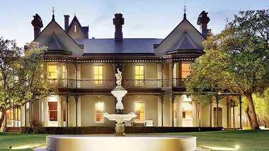 Elegant Victorian mansion with ornate details, large windows, and a central fountain, illuminated at dusk.