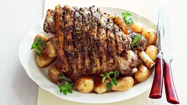 Roasted leg of lamb with rosemary and potatoes garnished with herbs on a white plate, accompanied by a knife and fork.