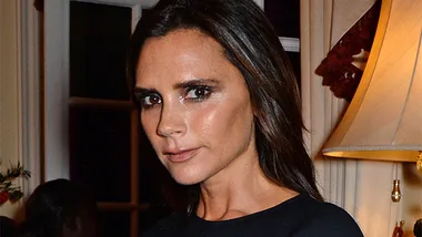 Victoria Beckham in a black dress at a formal event with elegant lighting in the background.