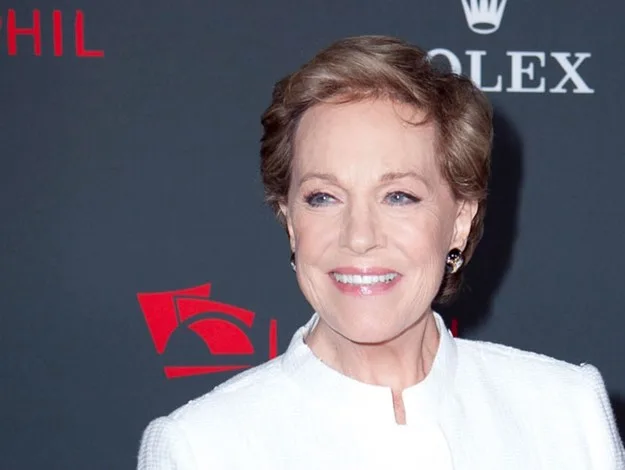 Woman in a white outfit smiling on a red carpet with logos in the background.