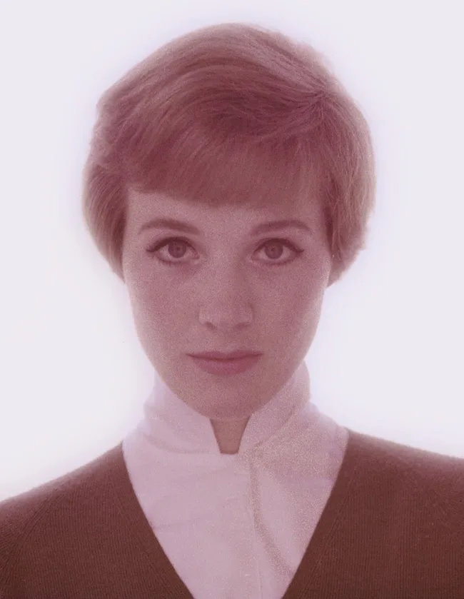 A person with short hair, wearing a white collared shirt and a dark sweater, looking directly at the camera.