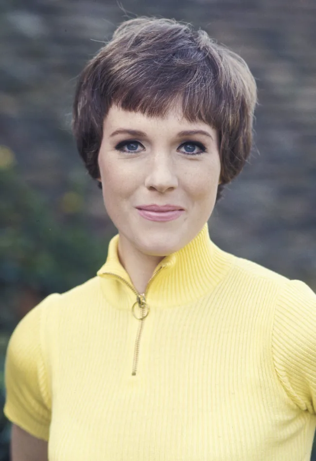 A woman with short hair wearing a yellow sweater, standing outdoors and smiling.