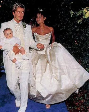A couple in formal wedding attire walks outdoors; the man carries a baby dressed in white.