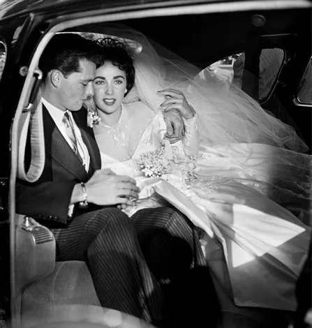 A bride in a wedding dress and veil sits in a car with a groom in a suit, both looking ahead.