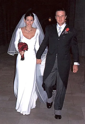 A bride in a white gown with a veil holds a red bouquet, walking hand-in-hand with a groom in a black suit and tie.