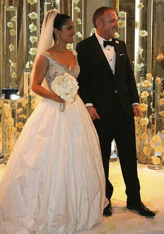 A bride in a white gown with a bouquet stands with a groom in a tuxedo amidst a decorative floral setting.