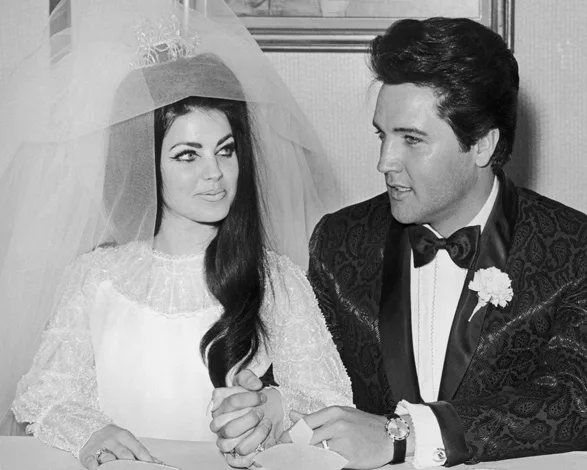 A bride in a white dress and veil holds hands with a groom in a dark suit and bow tie, seated and smiling at each other.
