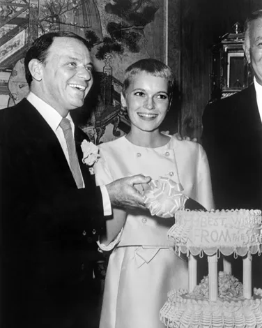 A couple smiling and cutting a tiered cake at a formal event; man in a suit, woman in a white dress with short hair.